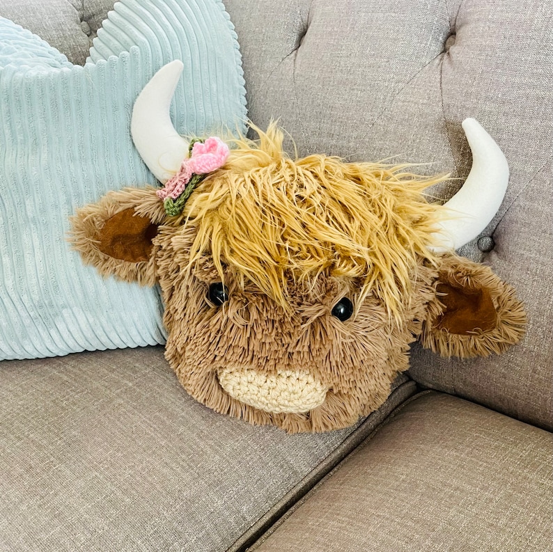 May include: A plush brown Highland Cow toy with a pink flower on its head. The toy is sitting on a gray couch.
