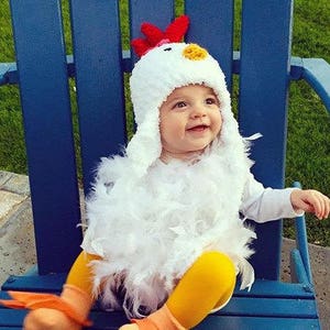 May include: A baby wearing a white chicken costume with a red comb and yellow beak. The costume is made of fluffy feathers and the baby is sitting on a blue chair.