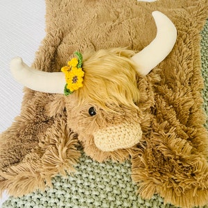 May include: A brown plush highland cow rug with a white horn and a yellow flower crown. The rug is on a green knitted blanket.