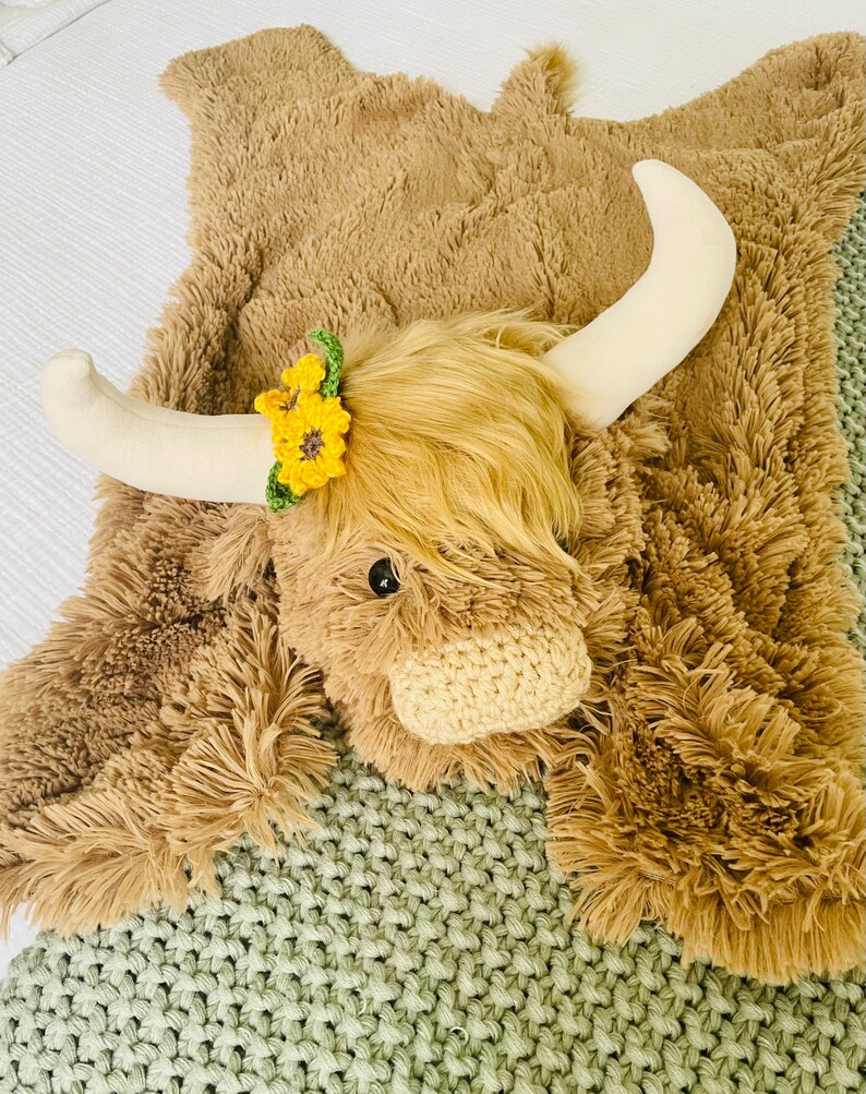 May include: A brown plush highland cow rug with a white horn and a yellow flower crown. The rug is on a green knitted blanket.