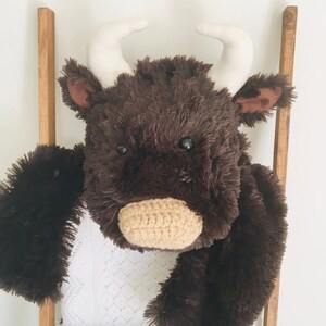 May include: A brown plush bull with white horns and a crocheted nose. The bull is sitting on a white blanket.