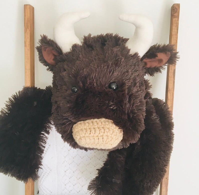May include: A brown plush bull with white horns and a crocheted nose. The bull is sitting on a white blanket.