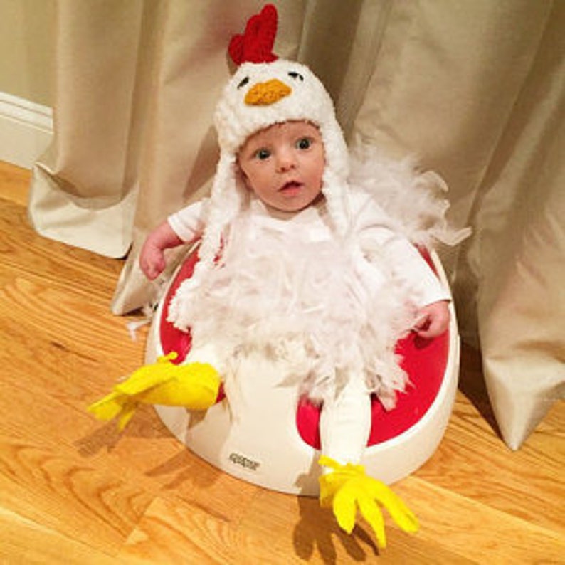 May include: A baby wearing a white chicken costume with yellow feet and a red and white crocheted hat. The costume is made of white feathers and the baby is sitting in a red and white baby seat.