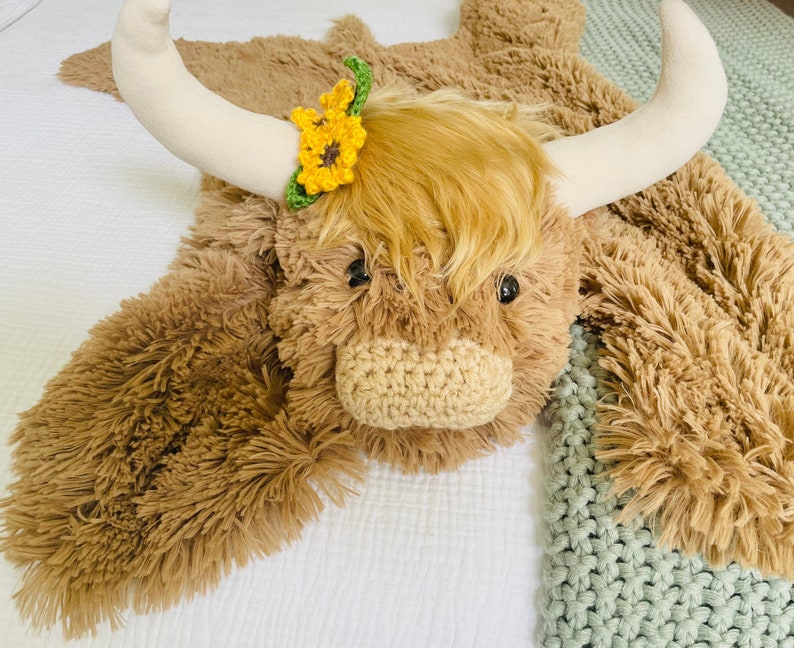 May include: A brown and white plush highland cow rug with a yellow flower hair bow. The rug is made of soft, fluffy fur and has a long, shaggy mane.