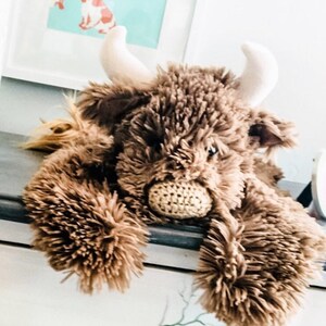 May include: A brown plush toy in the shape of a Highland cow with white horns. The toy is lying on a white surface with its head resting on the surface. The toy has a crocheted nose and a fluffy brown body.