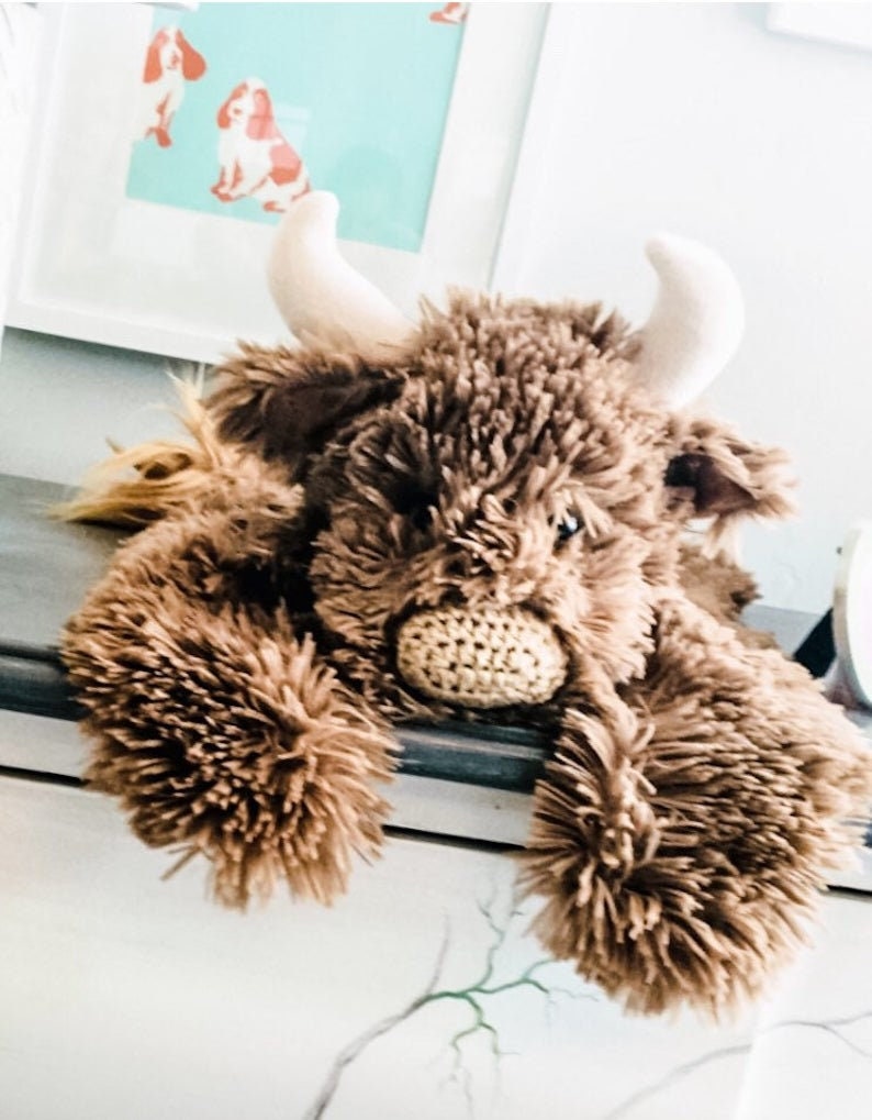 May include: A brown plush toy in the shape of a Highland cow with white horns. The toy is lying on a white surface with its head resting on the surface. The toy has a crocheted nose and a fluffy brown body.