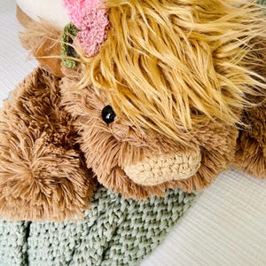 May include: A plush brown and white Highland cow toy with a pink flower crown, lying on a green knitted blanket.
