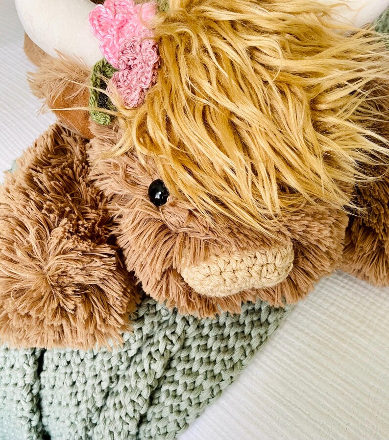 May include: A plush brown and white Highland cow toy with a pink flower crown, lying on a green knitted blanket.