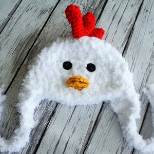 May include: White crocheted chicken hat with a red comb, yellow beak, and black eyes. The hat has long, fluffy ear flaps.