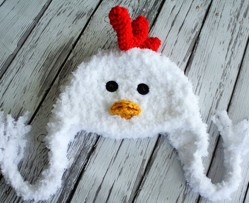 May include: White crocheted chicken hat with a red comb, yellow beak, and black eyes. The hat has long, fluffy ear flaps.
