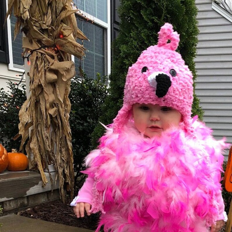 May include: A child dressed as a flamingo for Halloween. The child is wearing a pink crocheted hat with a black beak and white eyes. The child is also wearing a pink and white feathered boa.