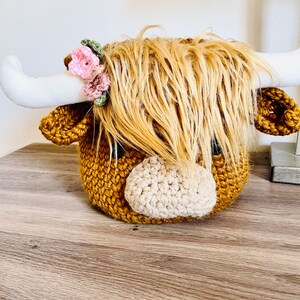 May include: A handcrafted crochet Highland cow head with long, shaggy, light brown yarn hair. The cow has white horns, a tan nose, and small pink and green floral accents. The body is a textured, golden-brown color.