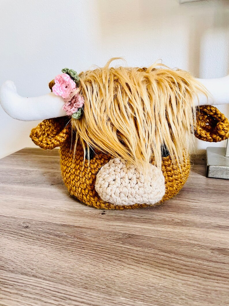 May include: A handcrafted crochet Highland cow head with long, shaggy, light brown yarn hair. The cow has white horns, a tan nose, and small pink and green floral accents. The body is a textured, golden-brown color.