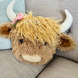 May include: A plush brown and white Highland cow head with a pink and green flower headband. The cow head is sitting on a gray couch.