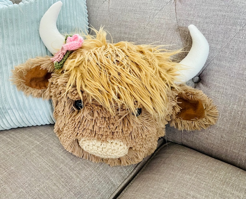 May include: A plush brown and white Highland cow head with a pink and green flower headband. The cow head is sitting on a gray couch.