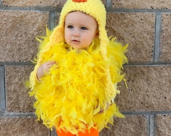 May include: A baby wearing a yellow chick costume with a yellow and orange hat. The costume is made of feathers and the hat has a red comb.