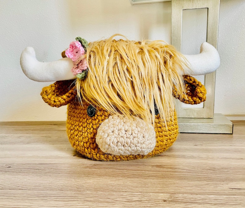 May include: A crocheted Highland cow head in shades of mustard yellow and beige. The cow has fluffy, light brown hair, white horns, and a small floral crown. The eyes are black buttons.