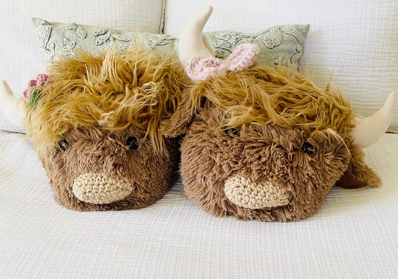 May include: Two brown plush Highland Cow head-shaped pillows with embroidered faces. One pillow has a pink bow on top.