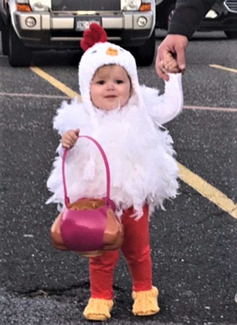 May include: A child dressed in a white chicken costume with red accents holds a pink and gold trick-or-treat bag. The child is wearing red pants and yellow shoes.