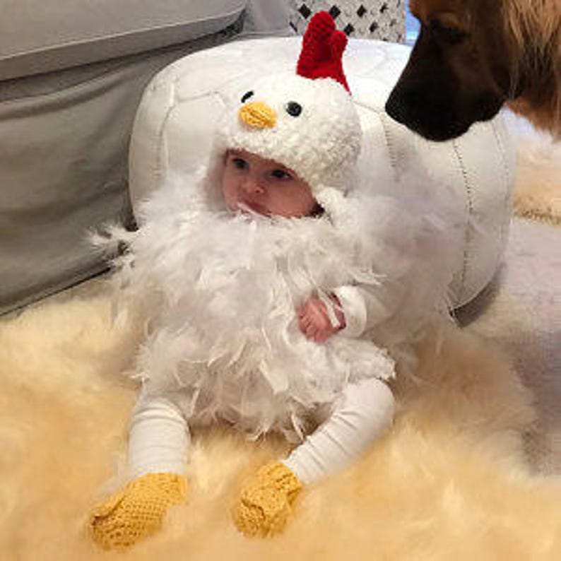 May include: A baby wearing a white chicken costume with red and yellow accents. The costume includes a hat, a feathery body, and booties.