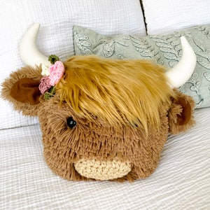 May include: A plush brown and white Highland cow head pillow with a pink and green flower crown. The pillow is sitting on a white bedspread.