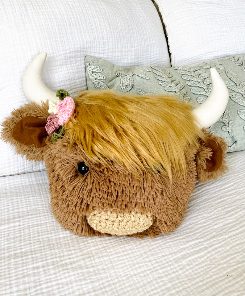 May include: A plush brown and white Highland cow head pillow with a pink and green flower crown. The pillow is sitting on a white bedspread.