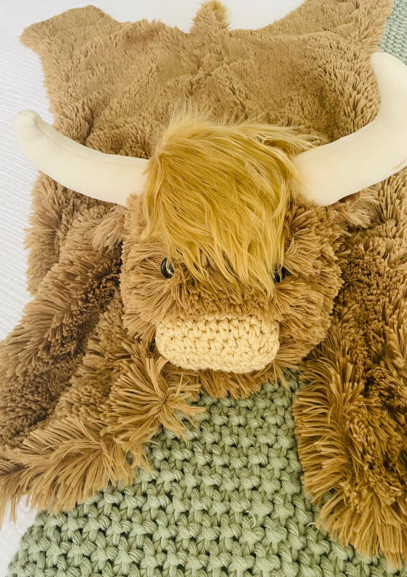 May include: A plush brown and white Highland cow rug with a crocheted nose and large, fluffy fur. The rug is lying on a green knitted blanket.