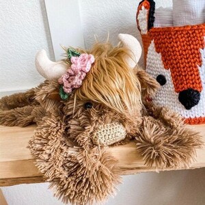 May include: A brown and white plush highland cow toy with a pink and green flower crown. The toy is lying on a wooden surface.