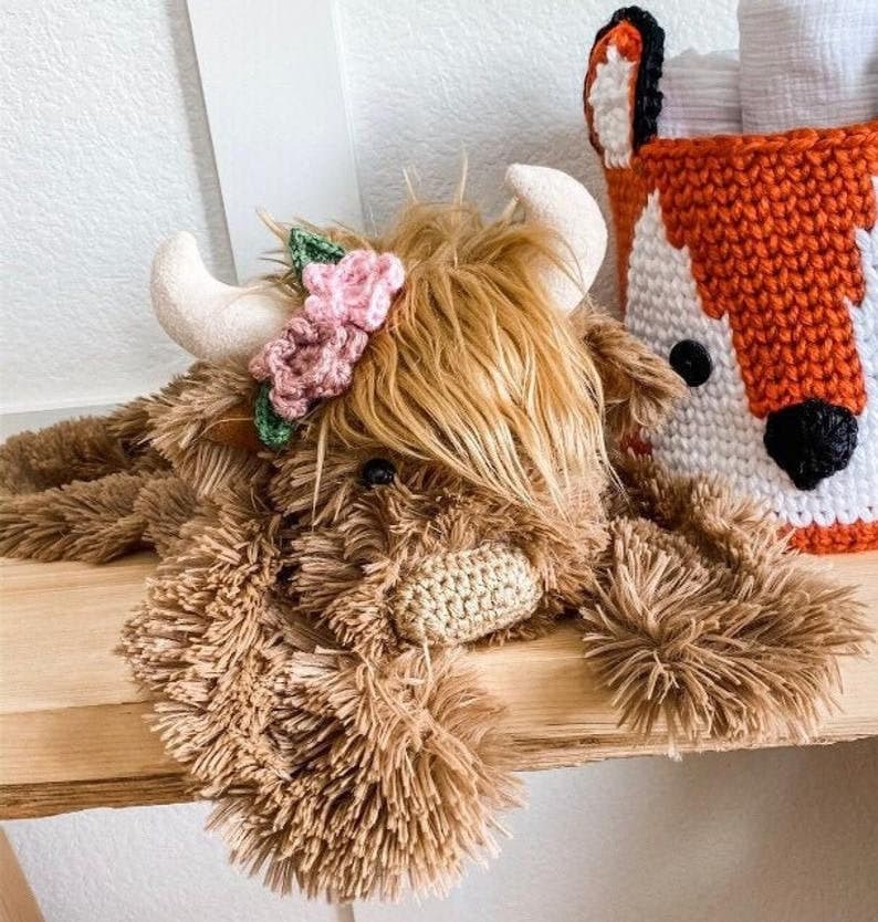 May include: A brown and white plush highland cow toy with a pink and green flower crown. The toy is lying on a wooden surface.