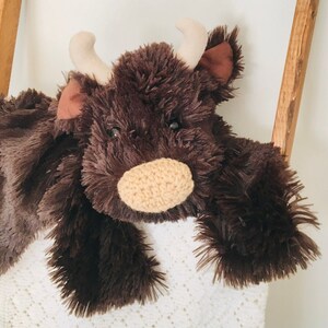 May include: A brown plush toy cow with a white blanket. The cow has a brown nose and large brown eyes.