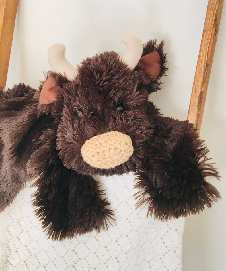 May include: A brown plush toy cow with a white blanket. The cow has a brown nose and large brown eyes.