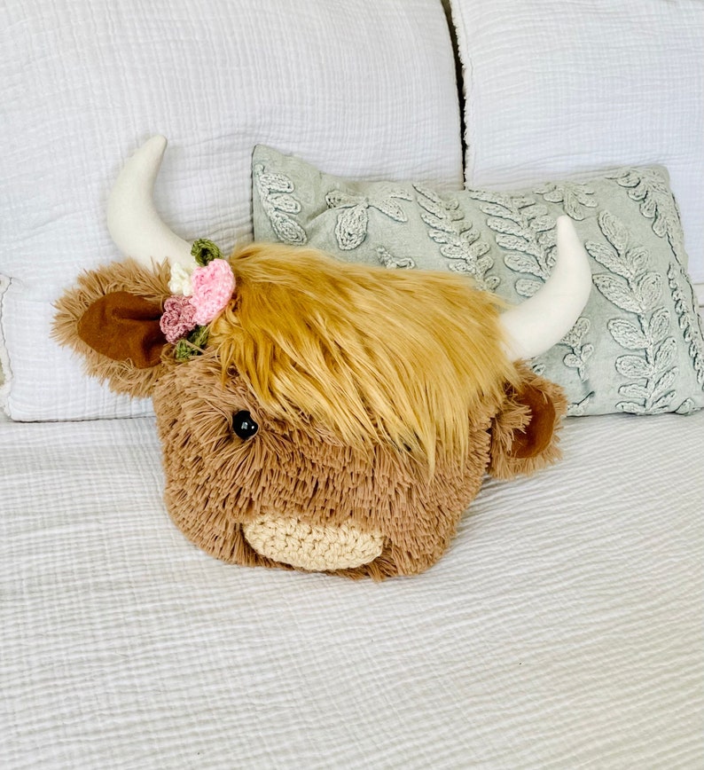 May include: A plush brown and white Highland cow head with a pink and green flower crown. The cow head is sitting on a white bedspread.