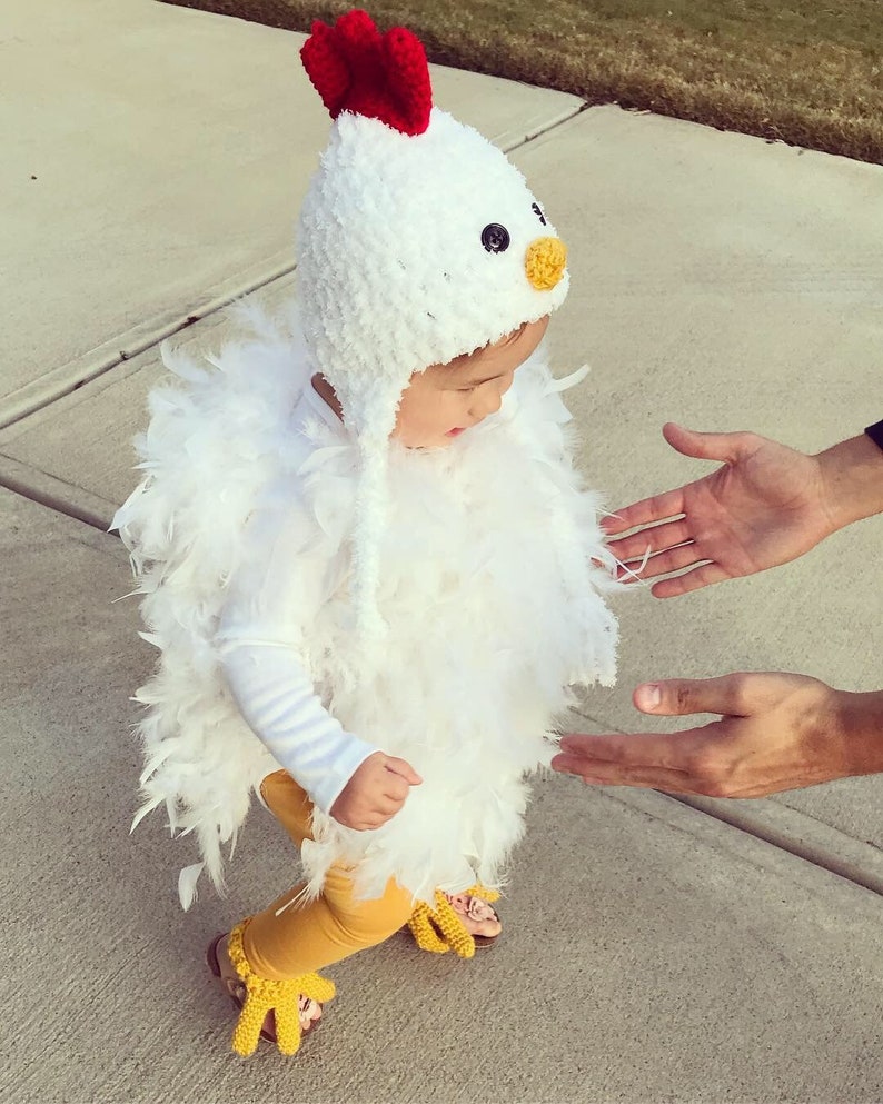 May include: A child wearing a white crocheted chicken costume with red and yellow accents. The costume includes a hat, a feathery body, and yellow crocheted feet.