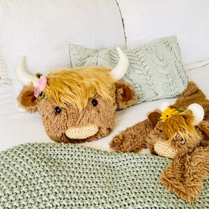 May include: Two plush brown and white Highland cow stuffed animals with floral headbands on a bed with a green knitted blanket.