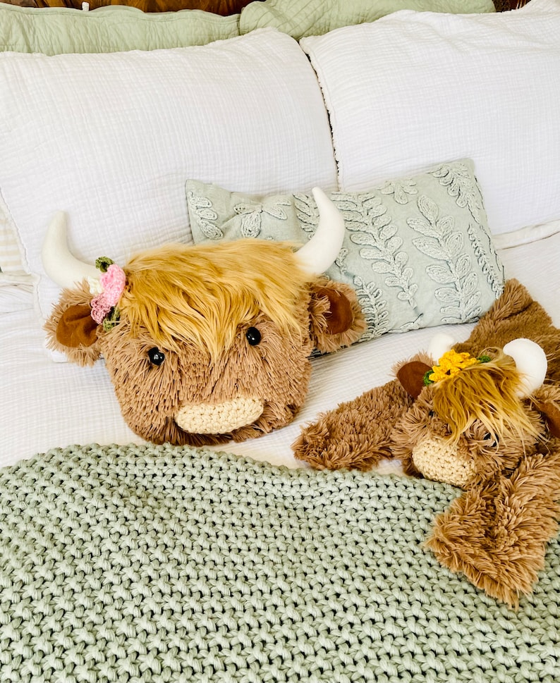 May include: Two plush brown and white Highland cow stuffed animals with floral headbands on a bed with a green knitted blanket.