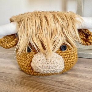 May include: A handcrafted, crocheted Highland cow head with a shaggy, light brown mane and white horns. The cow has button eyes and a light beige muzzle. The item is designed to be a decorative piece.