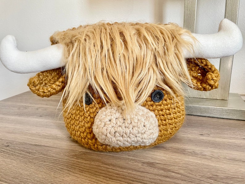 May include: A handcrafted, crocheted Highland cow head with a shaggy, light brown mane and white horns. The cow has button eyes and a light beige muzzle. The item is designed to be a decorative piece.