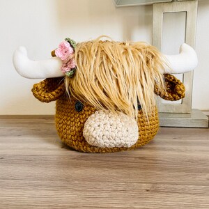 May include: A handcrafted crochet Highland cow head with a shaggy, light brown mane and a light tan muzzle. The cow has white horns and a small floral crown. The body is a dark brown color, and the eyes are black.