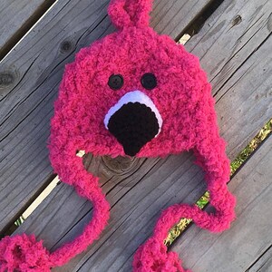 May include: Pink crocheted flamingo hat with black eyes and a black beak. The hat has long, fluffy pink ties.