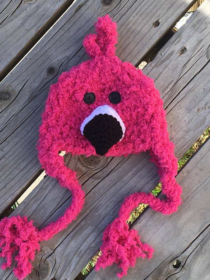 May include: Pink crocheted flamingo hat with black eyes and a black beak. The hat has long, fluffy pink ties.