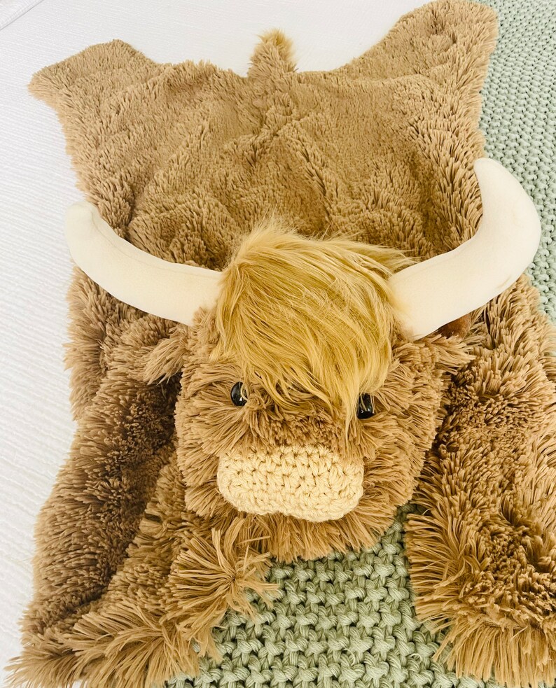 May include: A brown and white plush highland cow rug with a fluffy texture. The rug features a large head with a white nose and horns.