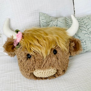 May include: A plush brown and white Highland cow head pillow with a pink and green flower headband. The pillow is lying on a white bedspread.