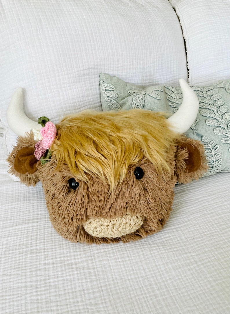 May include: A plush brown and white Highland cow head pillow with a pink and green flower headband. The pillow is lying on a white bedspread.