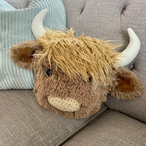 May include: A plush brown and white Highland cow head with a fluffy mane and a crocheted nose. The cow head is sitting on a gray couch.