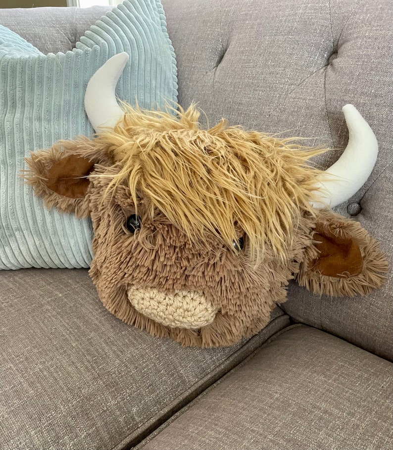 May include: A plush brown and white Highland cow head with a fluffy mane and a crocheted nose. The cow head is sitting on a gray couch.