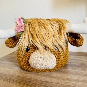 May include: A handcrafted, crochet Highland cow head with long, shaggy, light brown fur and white horns. The cow has a light tan muzzle and is adorned with a pink and green floral crown. The cow's head is a decorative item.