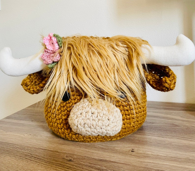May include: A handcrafted, crochet Highland cow head with long, shaggy, light brown fur and white horns. The cow has a light tan muzzle and is adorned with a pink and green floral crown. The cow's head is a decorative item.