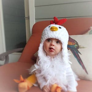 May include: A baby wearing a white crocheted chicken costume with a red comb and yellow beak. The costume has a fluffy white feather trim. The baby is sitting on a red couch.