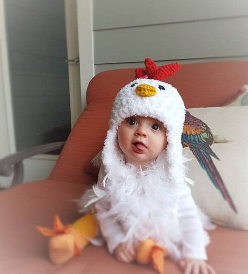 May include: A baby wearing a white crocheted chicken costume with a red comb and yellow beak. The costume has a fluffy white feather trim. The baby is sitting on a red couch.