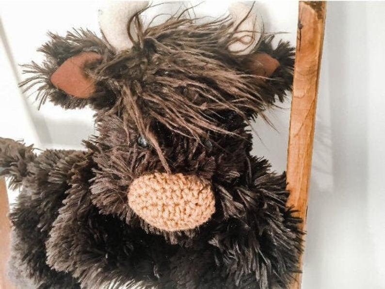 May include: A brown and black plush toy in the shape of a Highland cow with a crocheted nose and white horns.